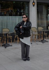 Kobieta w czarnej skórzanej kurtce, białym asymetrycznym topie, szarych spodniach i okularach przeciwsłonecznych stoi z założonymi rękami obok stolików i krzeseł kawiarnianych.