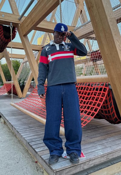 Polo de manga larga color marino con rayas rojas, combinado con jeans de mezclilla oscura de corte oversized. Gorro color marino con logo. Estructura de madera y red de color rojo en el fondo.