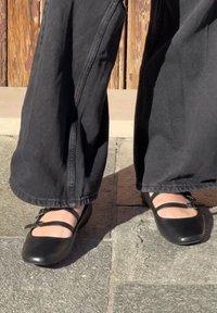 Melnas balerīnas ar divām plānām siksnām frontē, matēta apdare, noapaļota purngala, valkātas ar melnām platiem biksēm uz akmens virsmas.