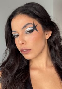 Närbild av ett ansikte med svarta stjärnformade sminkdetaljer runt höger öga. Silverögonskugga lyfter fram ögonlocket; läpparna är i en dämpad nude nyans.