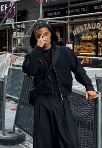 Sudadera negra con cremallera y capucha, sobre una camiseta negra, combinada con pantalones holgados negros con plisados. Se lleva un pequeño bolso negro acolchado en bandolera.