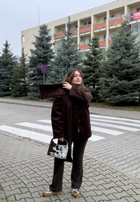 Bruine fleecejas met bijpassende sjaal, zwarte broek en een handtas met patroon. Op de achtergrond staan altijd groene bomen en een zebrapad met strepen.