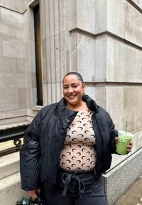 Femme en doudoune noire et haut à motif croissant tenant un smoothie vert devant un bâtiment en pierre, souriant à la caméra.