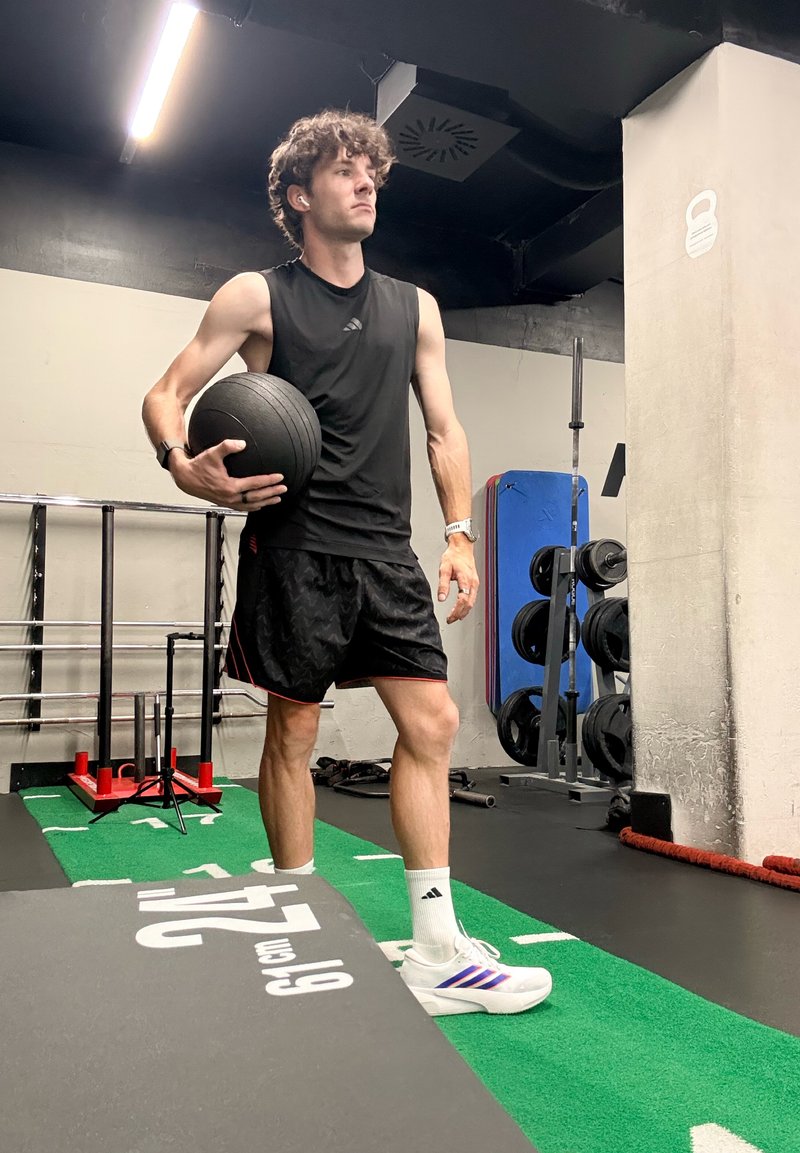 Jeune homme en tenue de sport noire tenant un ballon médicinal dans une salle de sport avec des poids, un sol rembourré et du matériel d'entraînement en arrière-plan.