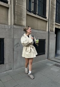 Trench-coat beige avec taille cintrée, sac à main noir, boisson verte, chaussettes blanches et baskets marron, debout sur un trottoir en pierre.
