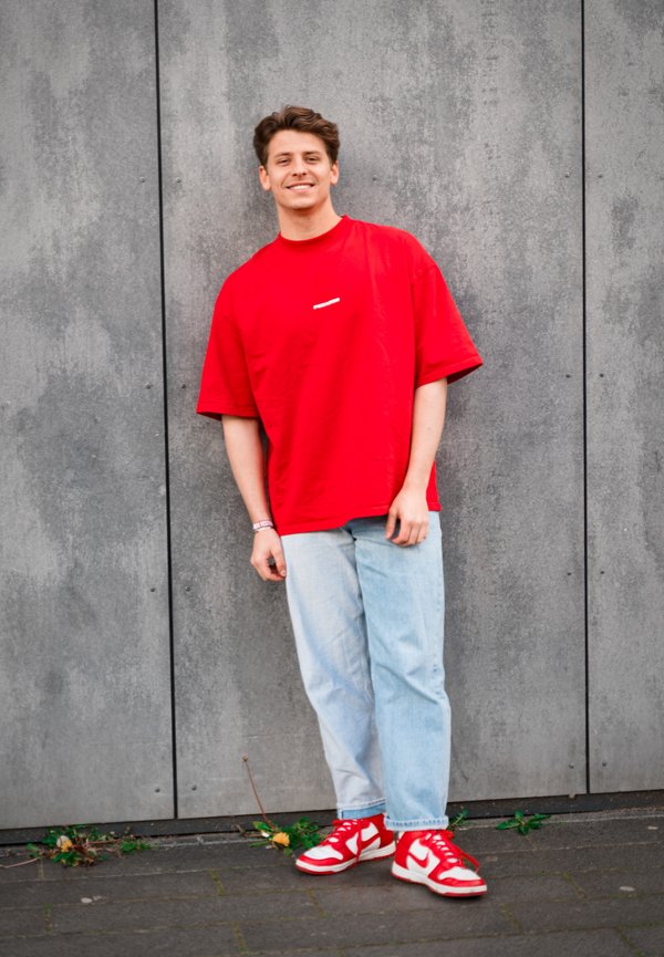 Camiseta roja de gran tamaño con logo blanco, vaqueros azul claros y zapatillas rojas con detalles blancos. Fondo de pared texturizada en gris.