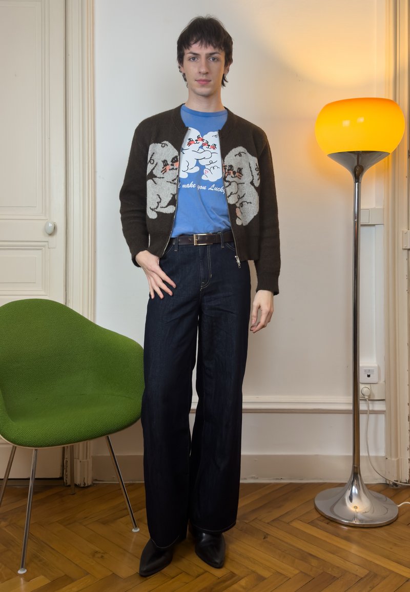 Young person stands indoors wearing a brown cardigan with puppy designs, blue graphic tee, dark wide-leg jeans, black shoes, near green chair and lamp.