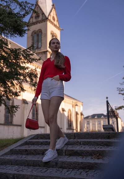 Suéter de punto rojo, pantalones cortos blancos y zapatillas blancas. Lleva un pequeño bolso rojo y sube escaleras de piedra con un edificio de fondo.
