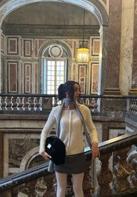 Femme en pull blanc, écharpe grise et jupe, debout sur un escalier en marbre orné à l'intérieur d'un bâtiment historique avec une grande fenêtre et une lanterne suspendue.