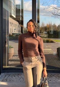 Brauner, gerippter Rollkragenpullover kombiniert mit hellgrauer, maßgeschneiderter Hose; hält eine dunkle Handtasche, mit Glasgebäude im Hintergrund und angelegtem Garten.