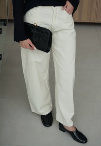 Άτομο που φοράει λευκό παντελόνι με φαρδιά μπατζάκια και μαύρα παπούτσια κρατάει μαύρο υφασμάτινο φάκελο με χρυσό κούμπωμα σε έναν μινιμαλιστικό εσωτερικό χώρο.