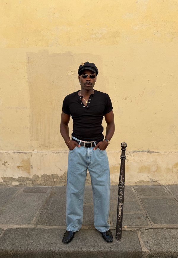 Homme en chemise noire, jean bleu clair, chapeau noir et lunettes de soleil, debout sur le trottoir contre un mur jaune décoloré avec un poteau métallique à proximité.