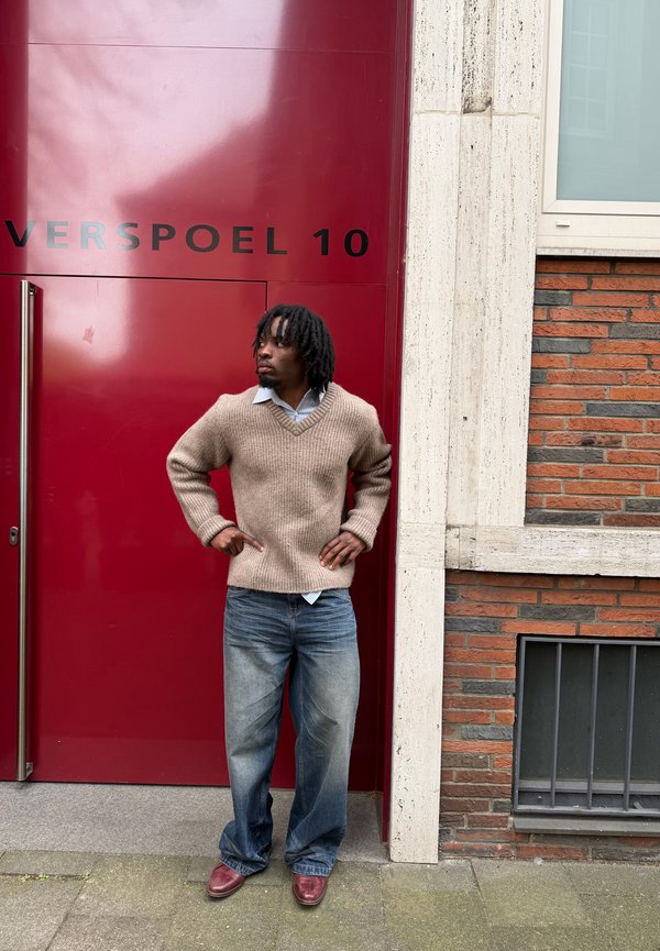 Mann in beigem Pullover und Jeans mit den Händen in den Hüften steht vor einer kastanienbraunen Tür mit der Aufschrift "VERSPOEL 10" neben einer Backsteinmauer.