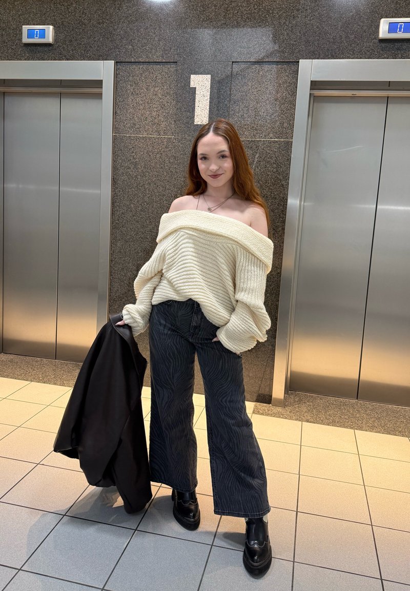 Jeune femme debout entre deux ascenseurs au 1er étage, portant un pull crème épaules dénudées, pantalon sombre à motifs, bottes noires, tenant une veste noire.