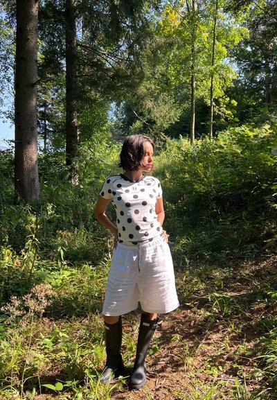 Camisa blanca con lunares negros y mangas cortas, pantalones cortos blancos y botas de goma negras hasta la rodilla en un entorno de bosque verde.