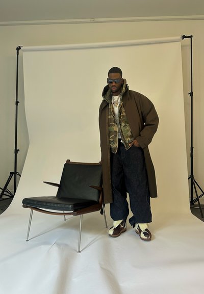 Hombre que lleva gafas de sol, abrigos en capas, pantalones anchos y botas únicas, de pie junto a una moderna silla de cuero negro y madera sobre un fondo blanco.