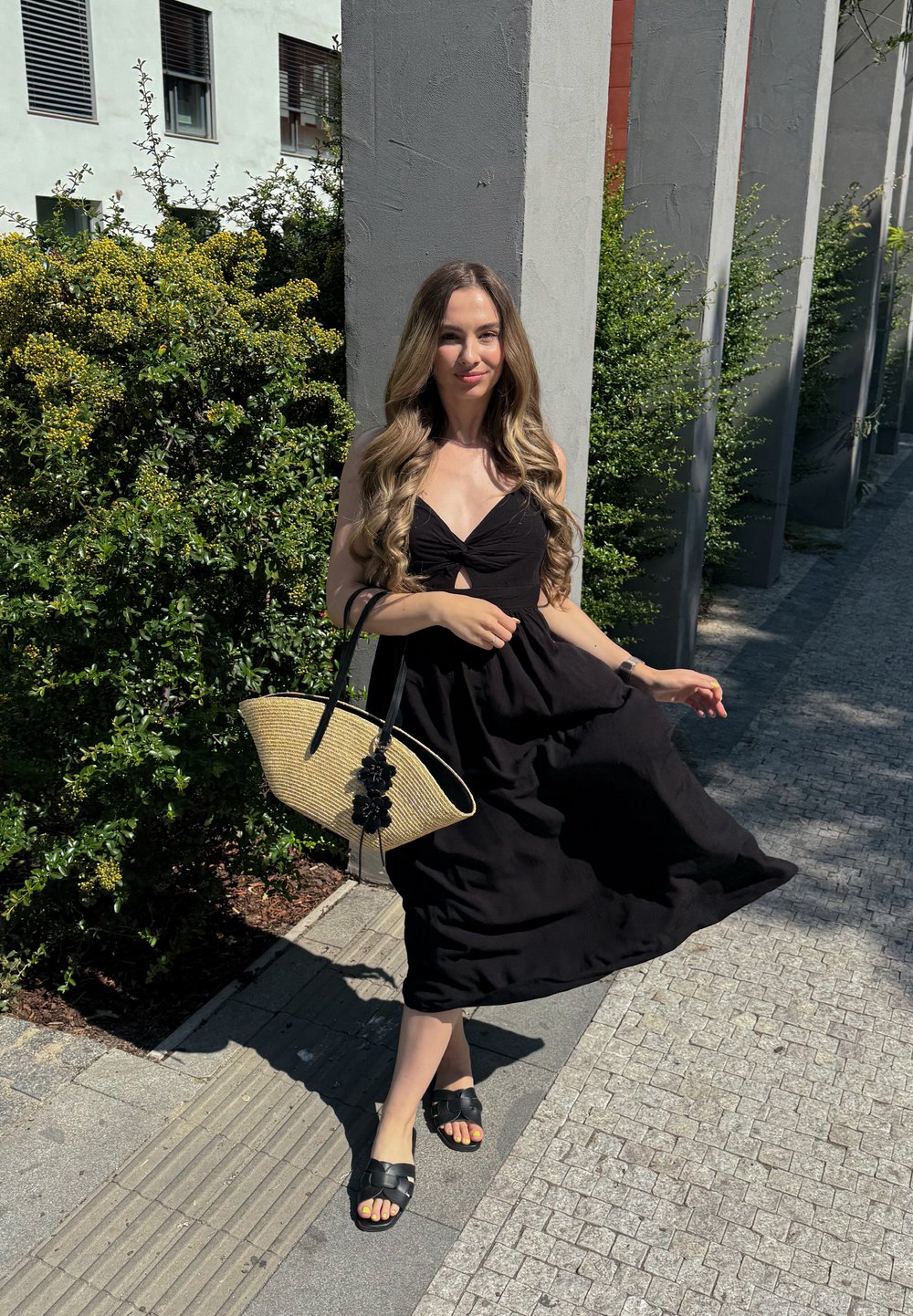 Femme en robe noire tenant un sac tissé, debout sur un chemin pavé avec des buissons verts et des piliers en béton sous un soleil éclatant.