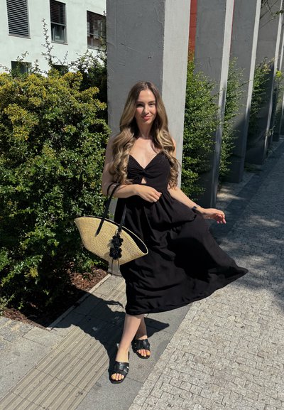Mujer con vestido negro sosteniendo un bolso tejido, de pie en un camino pavimentado con arbustos verdes y pilares de concreto bajo la luz brillante del sol.