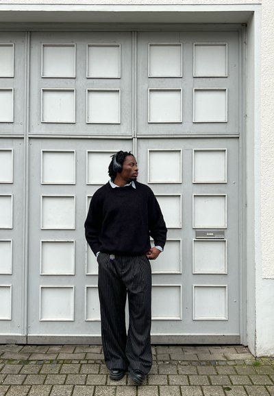Suéter negro de punto, pantalones de corte holgado a rayas con acento morado y auriculares, de pie junto a una puerta de panel gris.