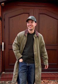 Uśmiechnięty mężczyzna w zielonej czapce bejsbolowej, oliwkowym płaszczu i ciemnym swetrze, w jeansach stoi z jedną ręką w kieszeni przed drewnianymi podwójnymi drzwiami.