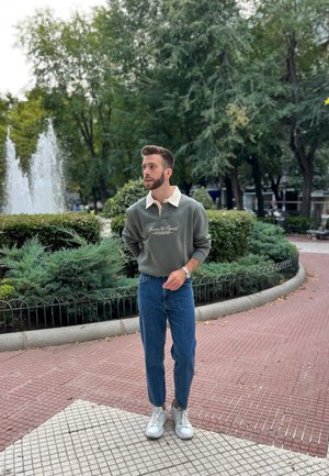 Sudadera verde con cuello blanco, con texto de logo, combinada con jeans azules y zapatillas blancas. Entorno al aire libre con vegetación.