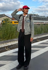 Džínsová bunda cez bielu košeľu a hnedú kravatu, doplnená širokými čiernymi nohavicami a čiernymi topánkami, s červenou šiltovkou a batohom.