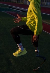 Atleta con chaqueta verde brillante, mallas negras y zapatillas de correr amarillas, utilizando una pierna protésica de alta tecnología mientras está en movimiento.