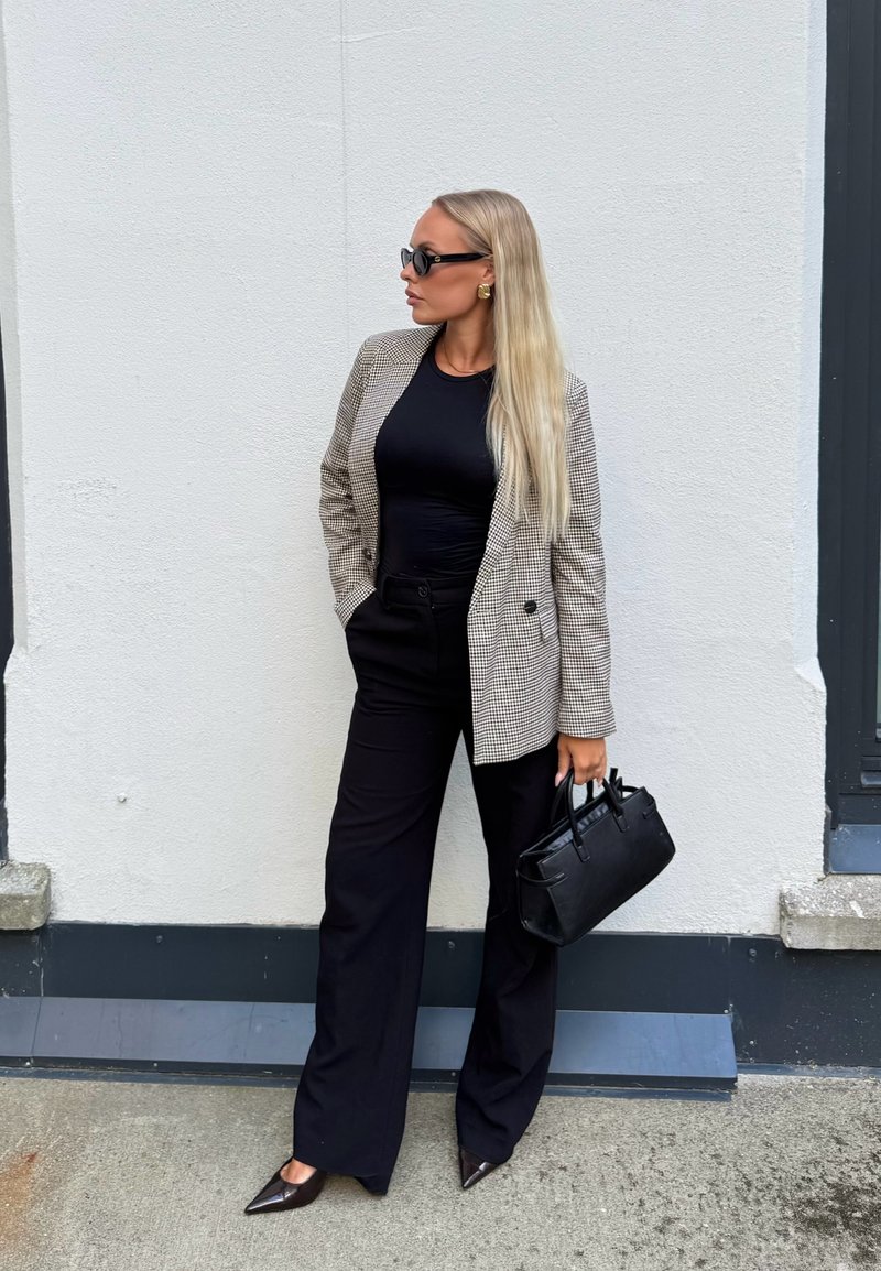 Femeie cu păr blond lung, care poartă un blazer în carouri, top negru, pantaloni evazați, pantofi ascuțiți, ochelari de soare și ține o geantă neagră.