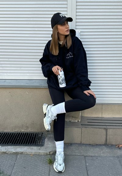 Sudadera negra de gran tamaño con un logo, leggings negros, calcetines blancos y zapatillas de deporte plateadas. Sosteniendo una botella de agua con un patrón; de pie contra una pared.