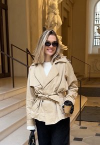 Femme en veste beige ceinturée et gants noirs, tenant une tasse de café et un sac à main, debout à l'intérieur d'un bâtiment avec des escaliers en marbre et une statue en arrière-plan.