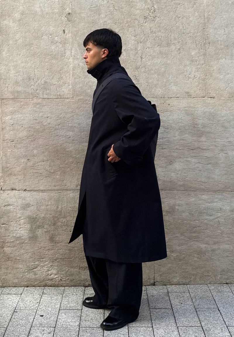 Cappotto nero con colletto alto, maniche lunghe e tasche laterali, abbinato a pantaloni neri larghi e scarpe lucide, posizionato di fronte a un muro strutturato.