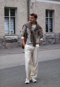 Man wearing a white and navy striped jumper, beige trousers, and trainers, with a brown bag and a knitted brown scarf draped over his shoulders.