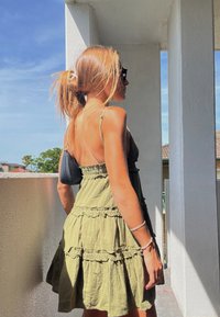 Femme aux cheveux châtain clair en queue de cheval, portant des lunettes de soleil et une robe verte sans manches, debout sur un balcon sous le soleil.