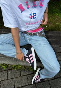 Weißes курzes Nike-T-Shirt mit rosa und blauer Grafik, hellblaue Jeans, schwarze Schuhe mit pinken Streifen, verzieller schwarzer Gürtel, weiße Ohrhörer.