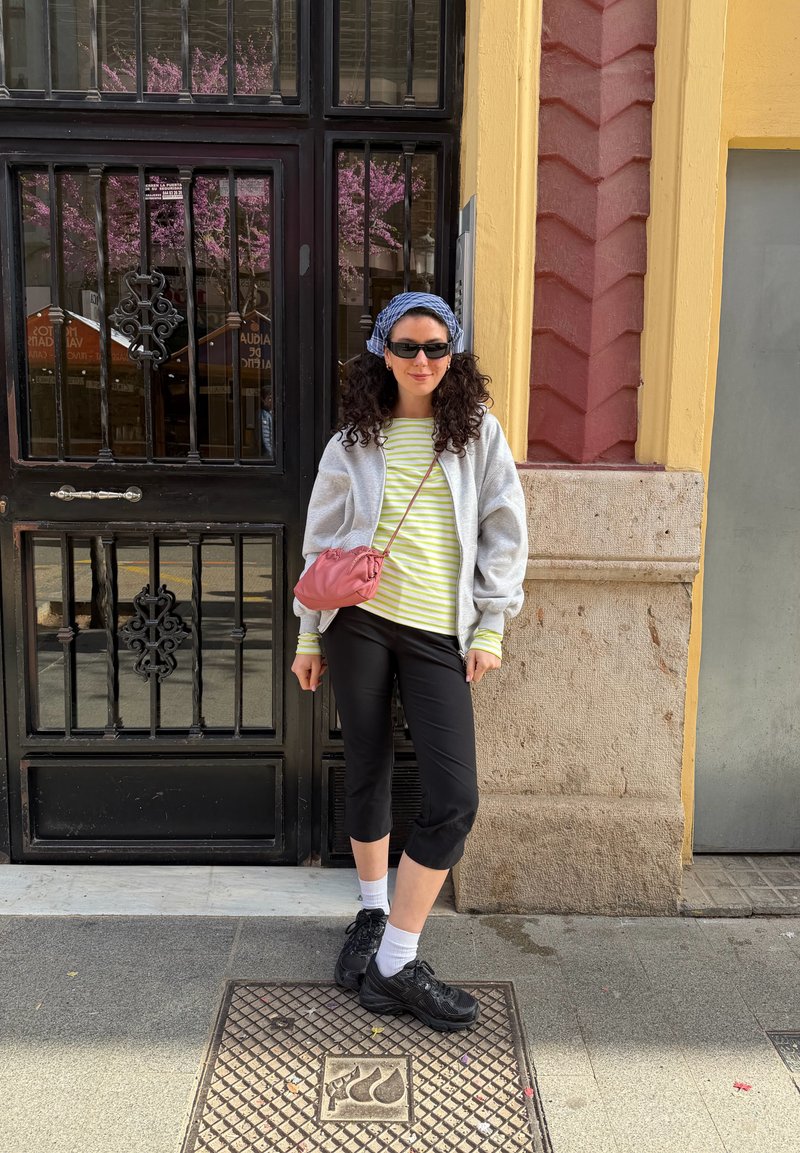 Femme aux cheveux bouclés, lunettes de soleil, foulard, chemise rayée, veste grise, pantalon capri noir, sac rose porté en bandoulière, debout près d’une porte métallique noire dans une rue de la ville.