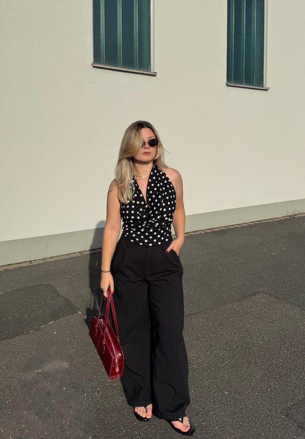 Femeie purtând pantaloni negri și un top halter negru cu buline, ținând o geantă roșie, stând pe trotuar lângă un perete bej cu două ferestre.