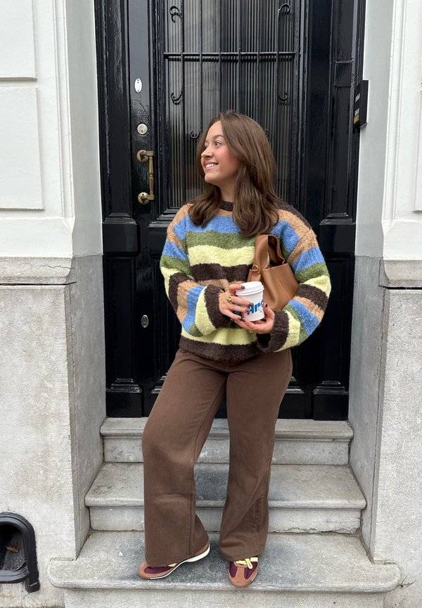 Farebný pruhovaný sveter so zelenými, modrými, žltými a hnedými odtieňmi; hnedé kordové nohavice; drží šálku kávy; stojí na kamenných schodoch.