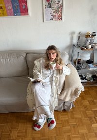 Personne en peignoir clair à motif floral, pyjama blanc et chaussons à thème cerise, tenant une tasse assise sur un canapé.