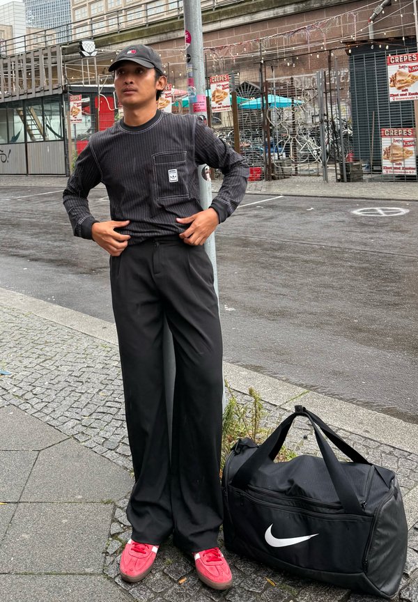 Camisa preta de manga longa com riscas e bolso, calças pretas, ténis vermelhos, mala de desporto preta da Nike, de pé numa rua de calçada.