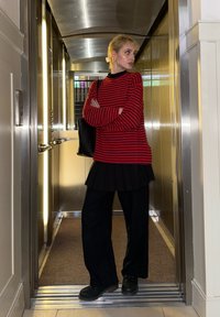 Jeune femme en pull rayé rouge et noir, jupe noire et pantalon, debout les bras croisés dans un ascenseur.