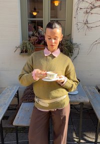 Maglione verde lavorato a maglia, camicia rosa a righe con colletto, pantaloni marroni a gamba larga, tiene una tazza da tè su un piattino bianco, ambiente esterno con fioriere.