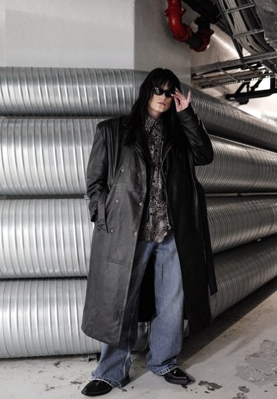Gabardina de cuero negro, camisa con estampado, jeans azules y zapatos negros. De pie frente a conductos metálicos en un entorno industrial.
