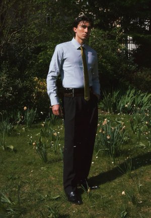 Hombre con camisa azul claro y corbata verde está de pie con una mano en el bolsillo en un jardín con césped, pequeñas flores blancas y arbustos frondosos.
