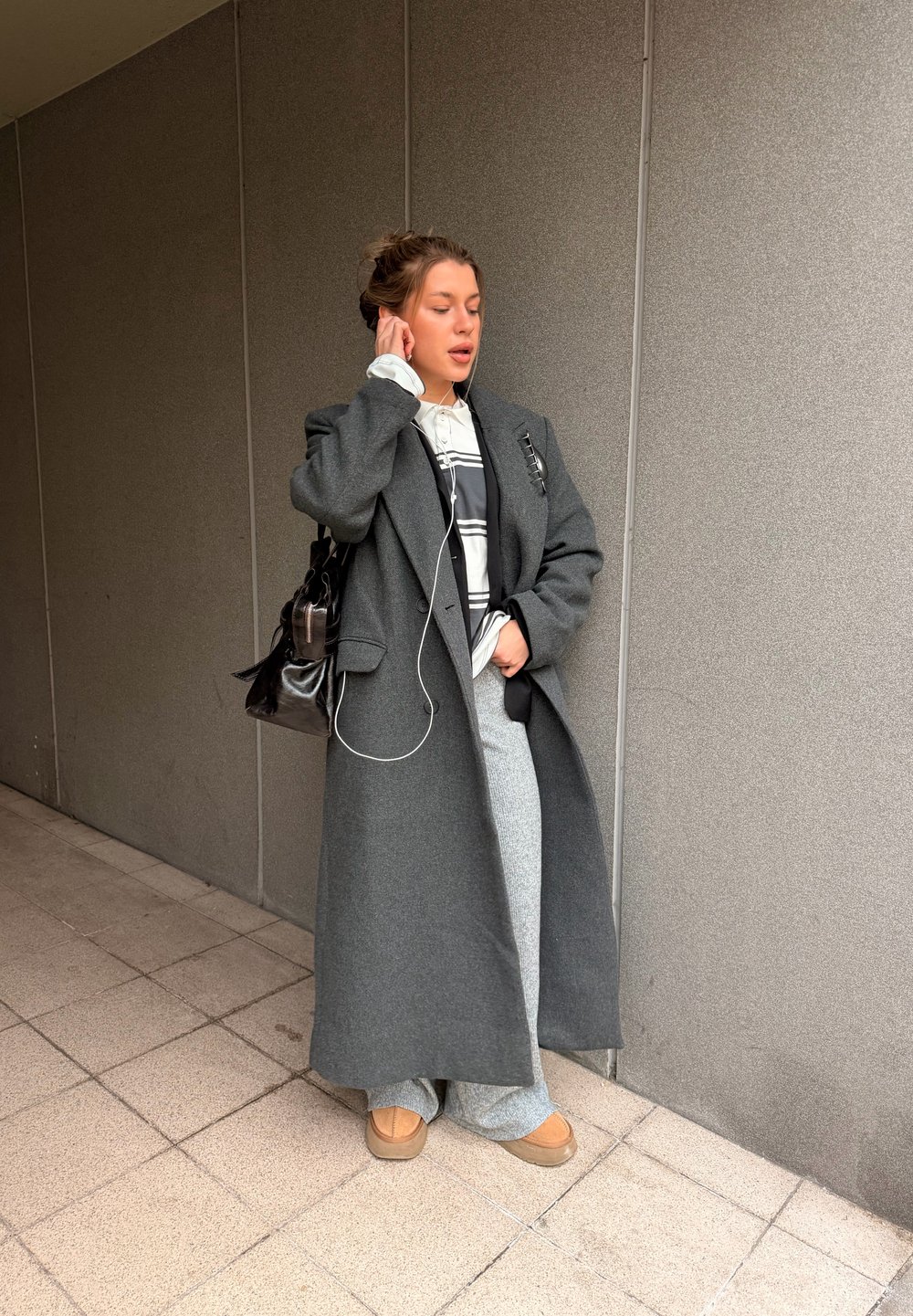 Mladá žena v dlhom sivom plášti, sivých nohaviciach a hnedých topánkach si rukou upravuje slúchadlo na ucho, stojí pri šedej obkladovej stene vo vnútri budovy.