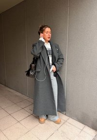 Mladá žena v dlhom sivom plášti, sivých nohaviciach a hnedých topánkach si rukou upravuje slúchadlo na ucho, stojí pri šedej obkladovej stene vo vnútri budovy.