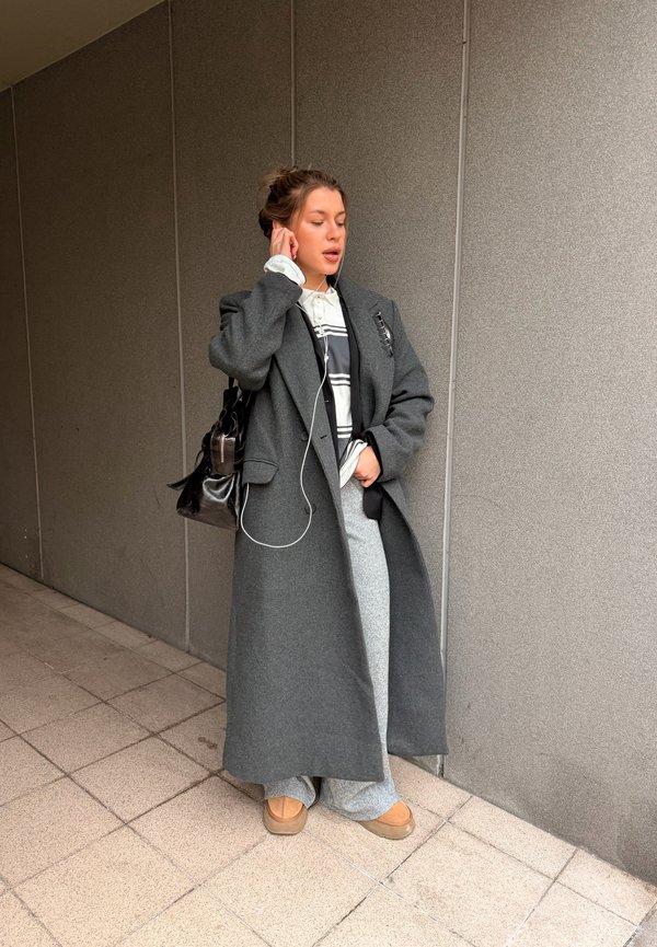 Mladá žena v dlhom sivom plášti, sivých nohaviciach a hnedých topánkach si rukou upravuje slúchadlo na ucho, stojí pri šedej obkladovej stene vo vnútri budovy.