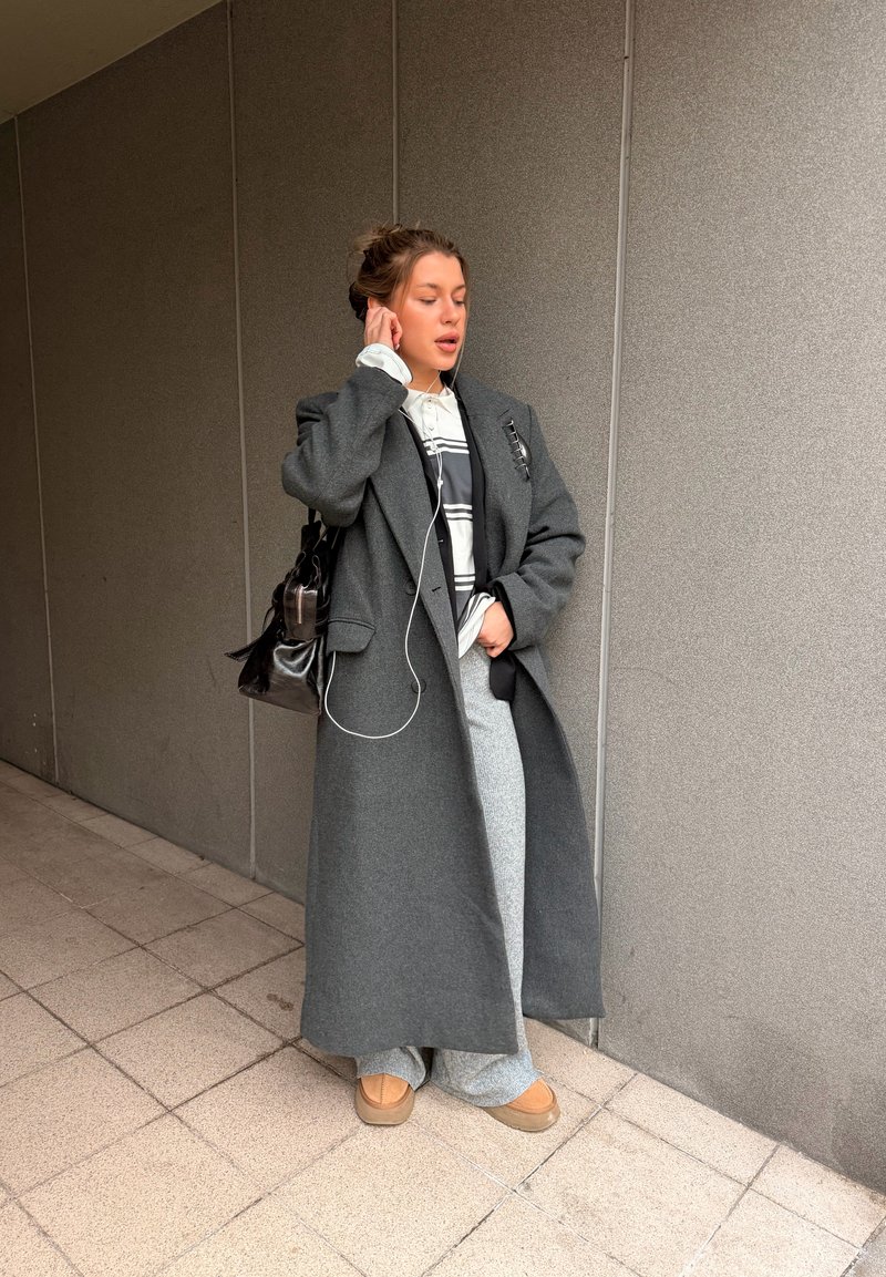 Giovane donna con lungo cappotto grigio, pantaloni grigi e scarpe color cuoio che si sistema un auricolare con la mano, in piedi davanti a una parete piastrellata grigia all'interno.