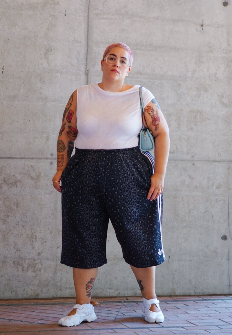 Shorts à imprimé léopard noir, chemise blanche, chaussures blanches. Les accessoires comprennent un sac bleu clair. Tatouages visibles sur les bras. Fond en béton.
