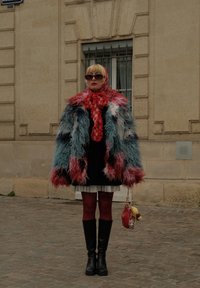 Kurtka z futerka w wielu kolorach z długimi rękawami, czerwony szalik, czarna sukienka, biała spódnica, czerwone rajstopy i czarne kozaki za kolano, trzymająca małą torebkę.