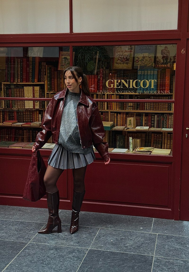 Casaco de couro bordeaux, suéter cinza de malha, saia cinza plissada, collants castanhos, botas de salto alto castanhas. Acompanham uma mala bordeaux combinando.